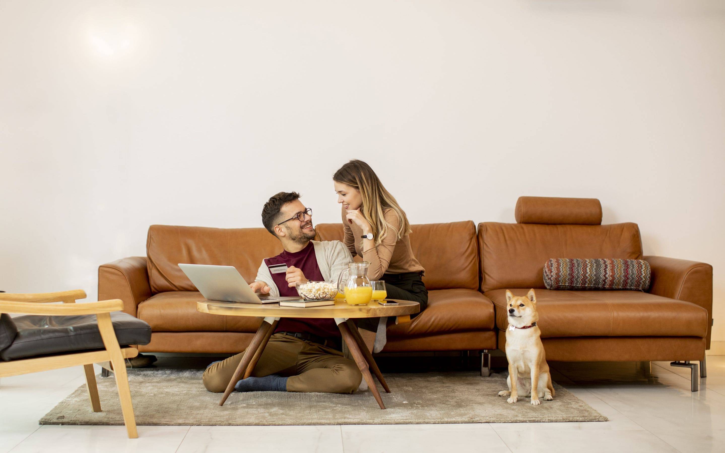 casal feliz na sala de estar, com seu cachorro. Um notebook sobre a mesa, pote com pipocas, suco de laranja.